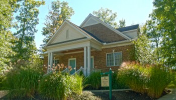 A small brick house with a porch, surrounded by lush greenery and tall grass, sits under a clear blue sky. A sign is visible in front.