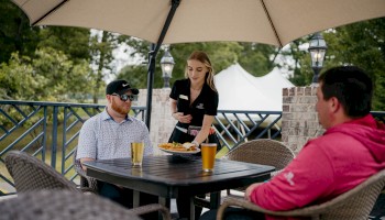 A waiter serves food to two people sitting under an umbrella at an outdoor table with drinks.