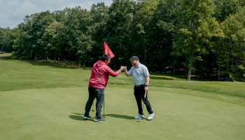 Two people on a golf course are giving each other a fist bump near a red flag.