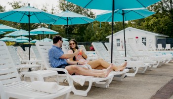 Two people relax on poolside loungers under blue umbrellas, holding drinks, surrounded by more empty loungers and rolled towels.