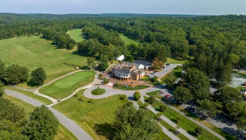 An aerial view of a golf course with a clubhouse surrounded by lush greenery and trees, featuring golf carts and a parking area.