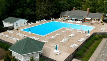 Aerial view of an outdoor swimming pool with surrounding lounge chairs, umbrellas, a gazebo, and nearby buildings in a green area.