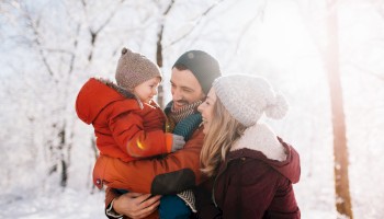 A family enjoying a snowy winter day, wearing warm clothing, smiling and embracing each other amidst snow-covered trees.