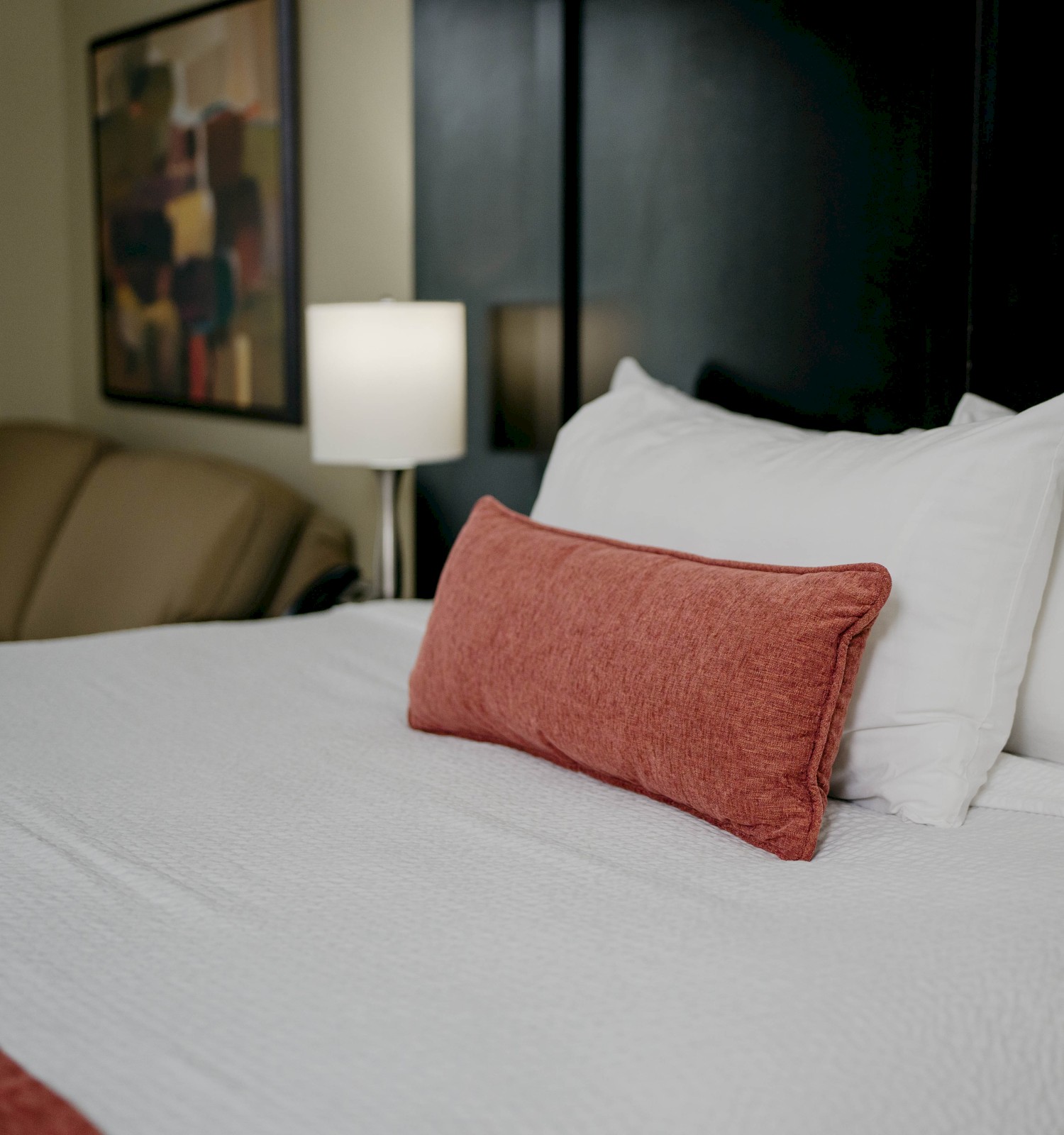 A neatly made bed with white linens and a red accent pillow, a couch, a lamp, and a painting in a cozy room setting.