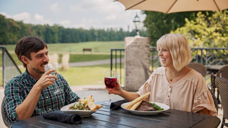 Two people are enjoying a meal and drinks on a patio with a scenic view. They are smiling and having a pleasant conversation.