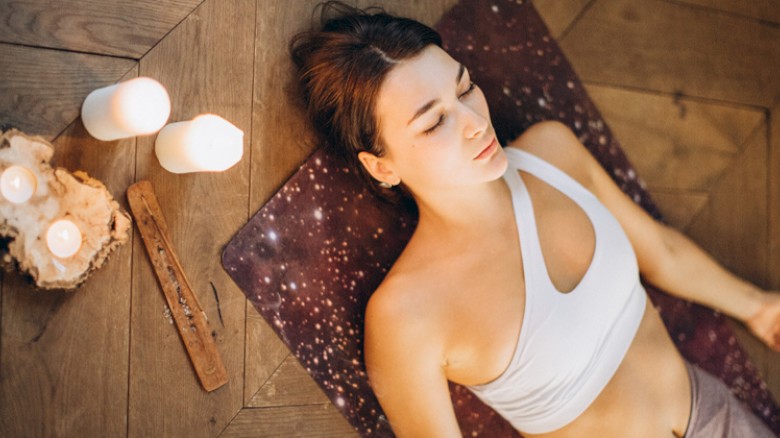 A person is lying on a yoga mat, practicing relaxation or meditation. Candles and incense are nearby, creating a calm atmosphere.