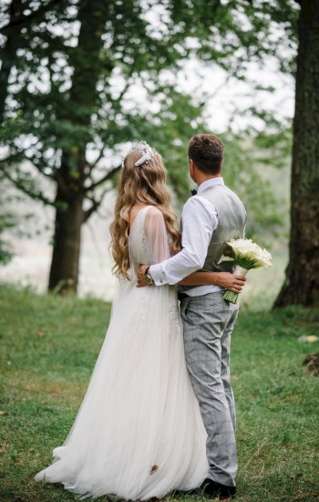 A bride and groom stand in a wooded area, embracing each other, with the bride holding a bouquet of flowers and facing away from the camera.