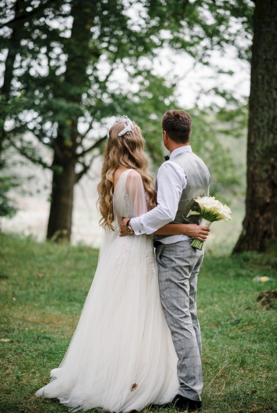 A bride and groom stand in a wooded area, embracing each other, with the bride holding a bouquet of flowers and facing away from the camera.