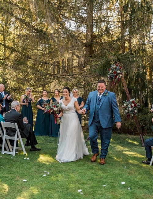 A couple is walking down the aisle outdoors, surrounded by guests applauding. The setting is a forested area with chairs for the ceremony.