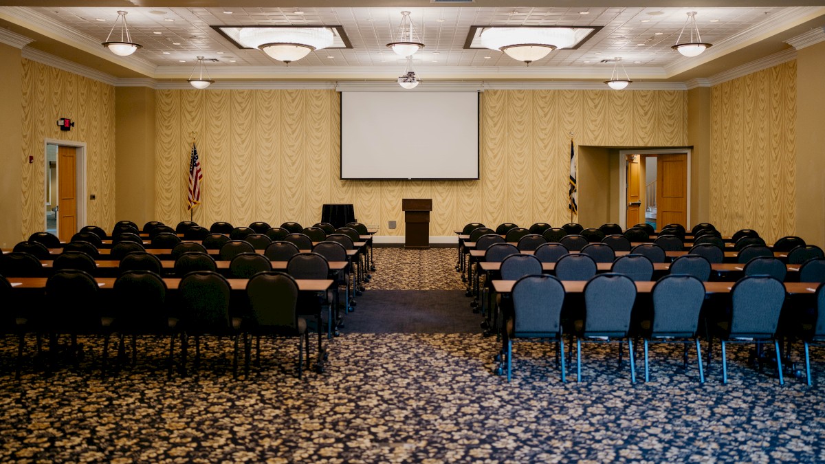 This image shows an empty conference room with rows of chairs and tables facing a podium, projection screen, and American flags.