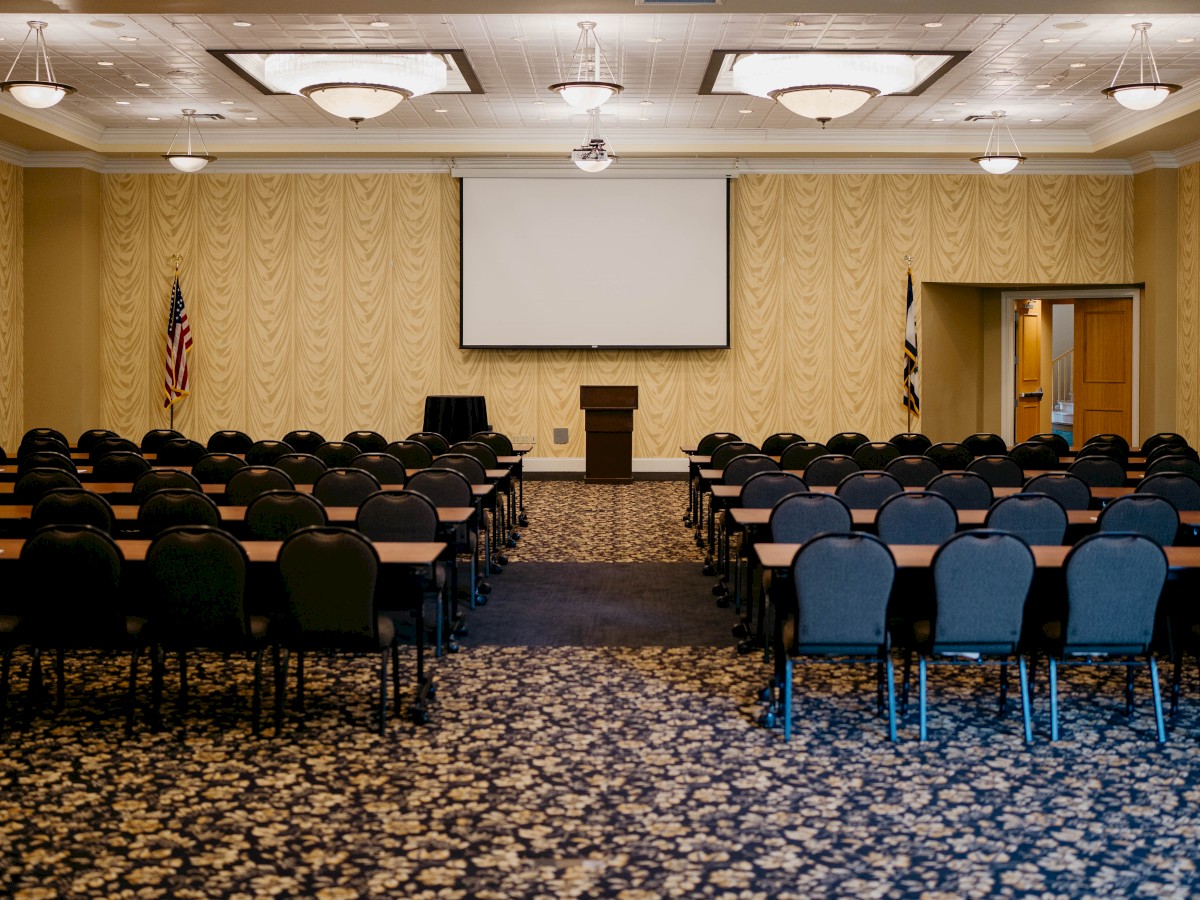 This image shows an empty conference room with rows of chairs and tables facing a podium, projection screen, and American flags.