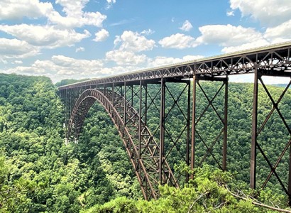 A large steel arch bridge spans a lush, green forest under a bright, blue sky with scattered clouds.