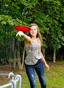 A person is playing disc golf, aiming a red disc towards a metal basket with chains, surrounded by green trees.