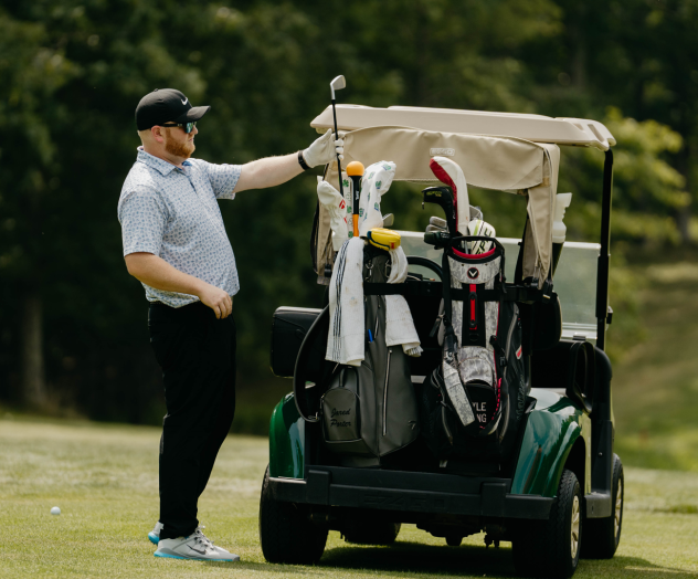 A person is standing by a golf cart on a grassy course, holding a golf club, with various clubs and equipment in the cart.