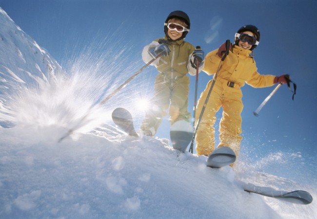 Two people in ski gear are skiing down a snowy slope under a clear blue sky, with snow spraying around them.