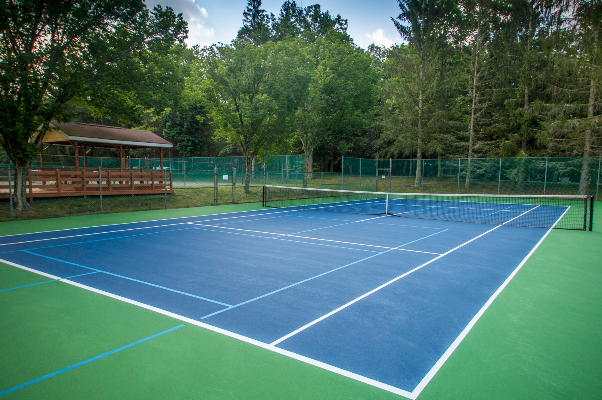 A tennis court with blue and green surfaces, surrounded by trees and a fence, with a wooden structure nearby.