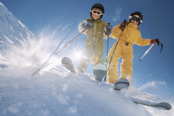 Two people in colorful ski gear are skiing downhill, kicking up snow under a clear blue sky.