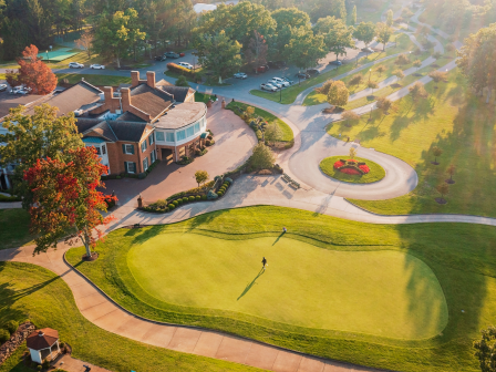 Aerial view of a golf course with a clubhouse, greenery, walking paths, and a person on the putting green during sunset.