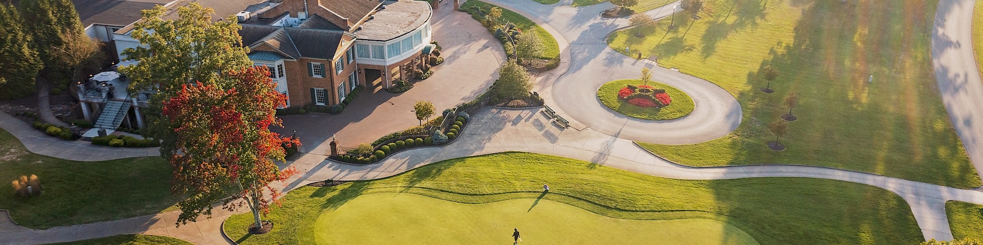 The image shows an aerial view of a golf course with a clubhouse, green, pathways, and surrounding trees in sunlight.