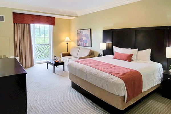 A hotel room with a king-sized bed, sofa, and TV. It features lamps, a table, and a window with drawn curtains, creating a cozy ambiance.