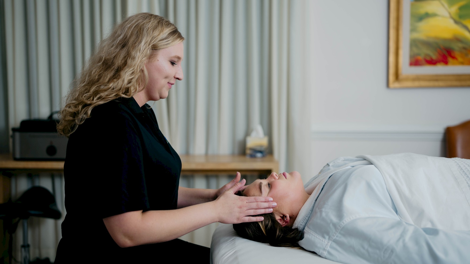 A person receives a facial massage from a therapist in a spa-like setting, surrounded by soft lighting and calming decor.