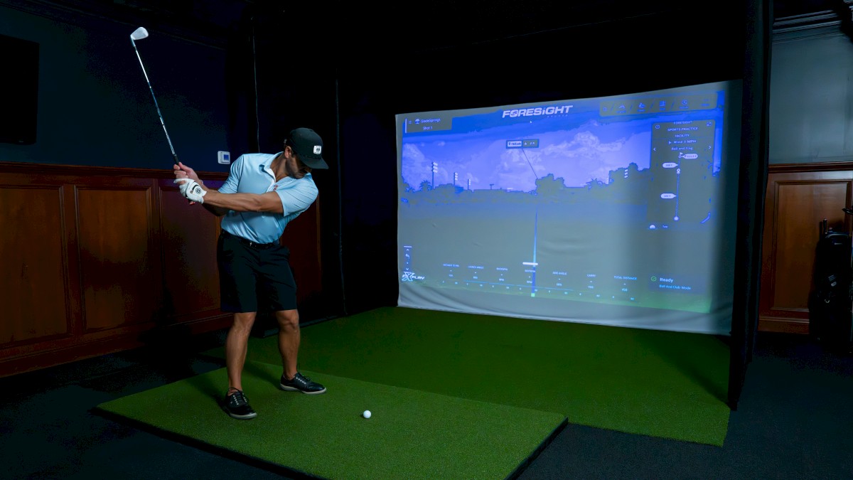 A person is playing golf indoors using a golf simulator, shown by the screen displaying a virtual golf course.