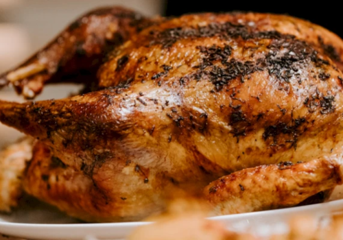 The image shows a roasted chicken with crispy, golden-brown skin, seasoned with herbs, served on a white plate.