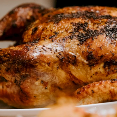 The image shows a roasted chicken with crispy, golden-brown skin, seasoned with herbs, served on a white plate.