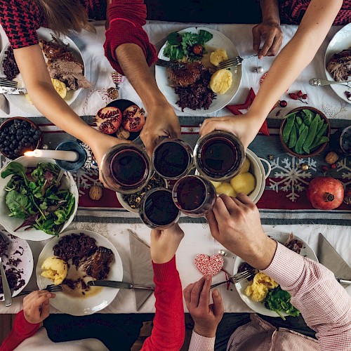 A festive table setting with plates of food, salads, and holiday decor. People raise glasses in a toast, celebrating together.