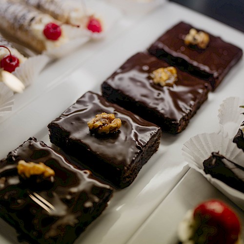 The image shows a selection of pastries on a platter, including chocolate brownies with walnuts, chocolate cakes with strawberries, and cannoli.