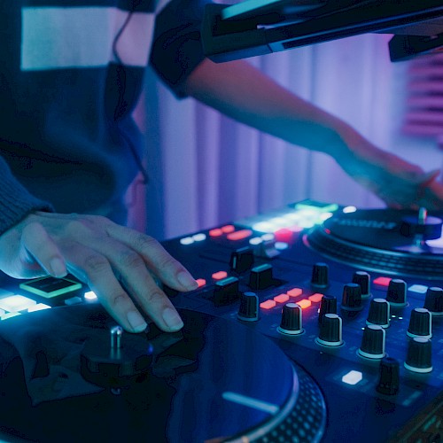 A DJ is mixing music on a console with glowing lights and controls in a dimly lit room, creating an energetic atmosphere.