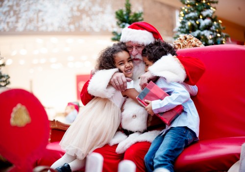 Santa hugging two children, one holding a gift, sitting on a red chair with festive decorations around them.