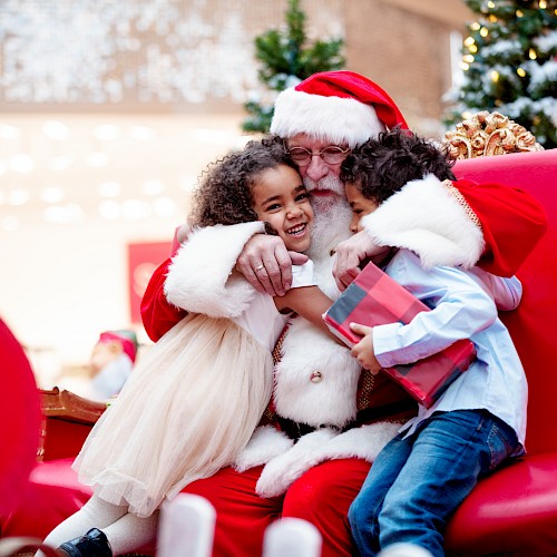 Santa hugging two children, one holding a gift, sitting on a red chair with festive decorations around them.