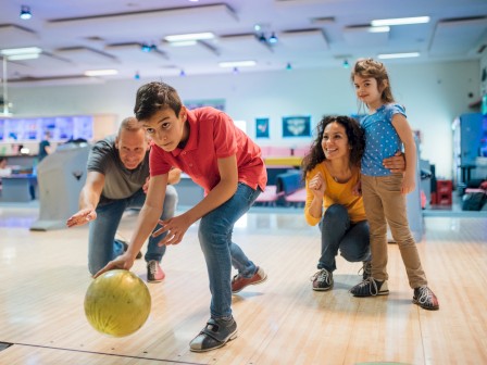 A family bowling together, with a young boy in a red shirt rolling a bowling ball, and three others watching and cheering.