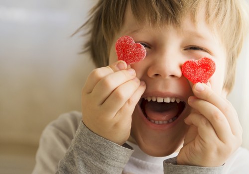 A child is smiling widely while holding two heart-shaped gummy candies in front of their eyes.