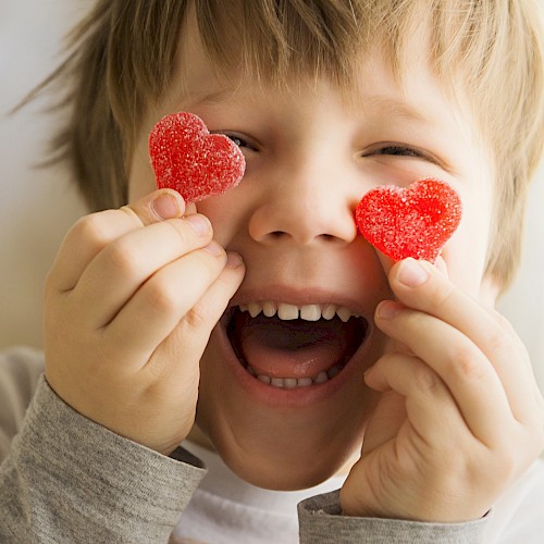 A child is smiling widely while holding two heart-shaped gummy candies in front of their eyes.