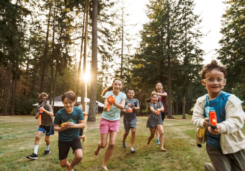 Children are playing with toy water guns outside, running and laughing in a forested area with the sun shining through the trees.