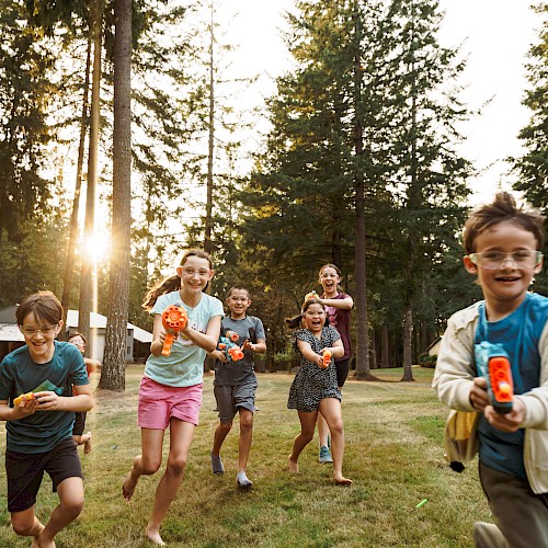 Children are playing with toy water guns outside, running and laughing in a forested area with the sun shining through the trees.