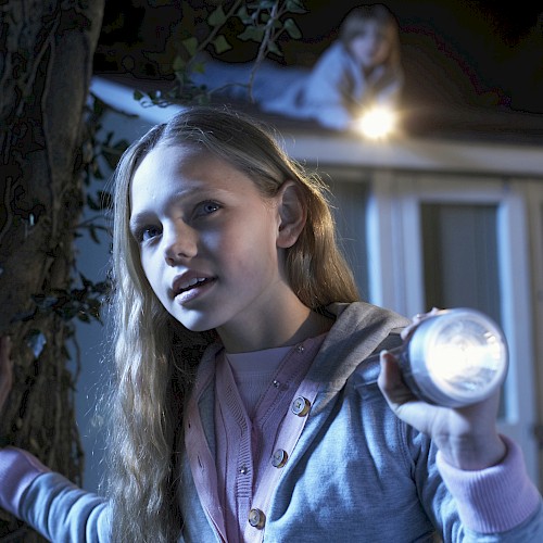 A girl holding a flashlight stands in front of a building at night, while another person is visible on the roof.