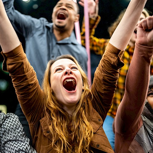 A diverse group of people are cheering energetically with their arms raised in a celebratory atmosphere.