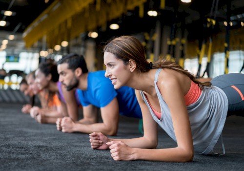 A group of people in a gym performing planks on a dark mat. They are aligned in a row, focused, and wearing athletic clothing.