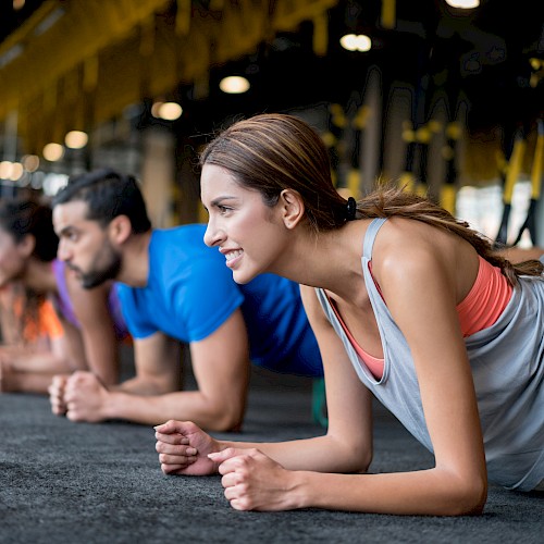 A group of people in a gym performing planks on a dark mat. They are aligned in a row, focused, and wearing athletic clothing.