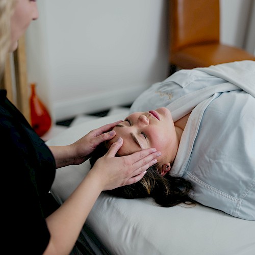 A person is receiving a facial massage while lying on a massage table, covered by a blanket in a serene room setting.