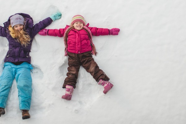 Two children in winter clothing making snow angels on fresh snow, smiling and enjoying a snowy day outdoors.