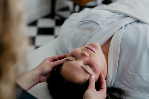 A person is receiving a facial massage while lying on a bed, with a therapist attending to them in a serene setting.