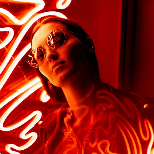 A person wearing sunglasses is illuminated by red neon lights with swirling patterns around them, creating an artistic and vivid scene.