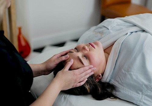 A person is receiving a facial massage while lying on a bed, covered with a light-colored sheet.