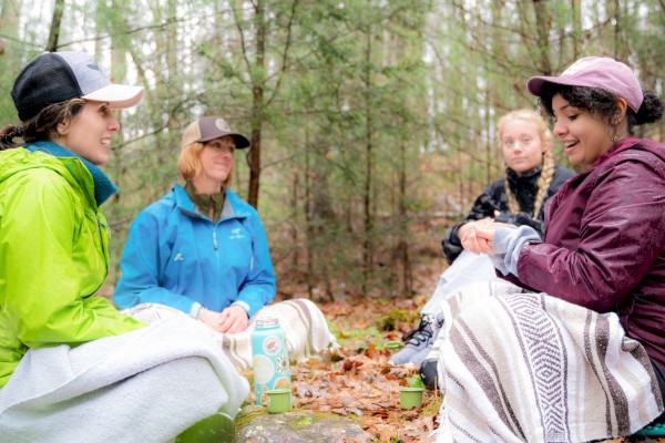 Four people sit outdoors on a forest floor, wearing jackets and caps, wrapped in blankets, with tea lights and a thermos nearby.